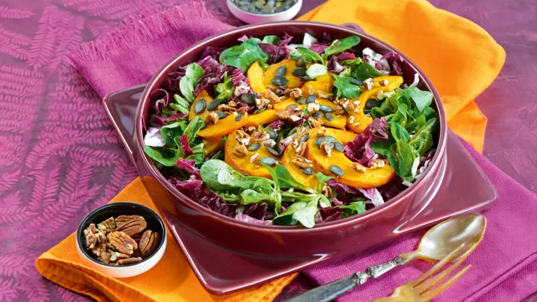 Blattsalat mit Radicchio, Feldsalat, gebratenen Kürbisscheiben, Pekannüssen und Kürbiskernen in einer dunkelroten Schale, serviert auf pinken und orangen Tüchern.
