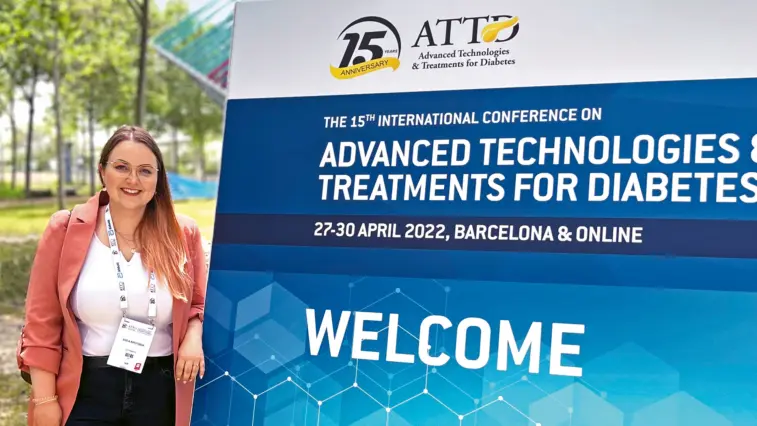 Autorin Svea Krutisch steht neben einem Aufsteller des ATTD-Kongresses. Darauf steht: „The 15th International Conference on Advanced Technologies & Treatments for Diabetes – 27-30 April 2022, Barcelona & online – Welcome“