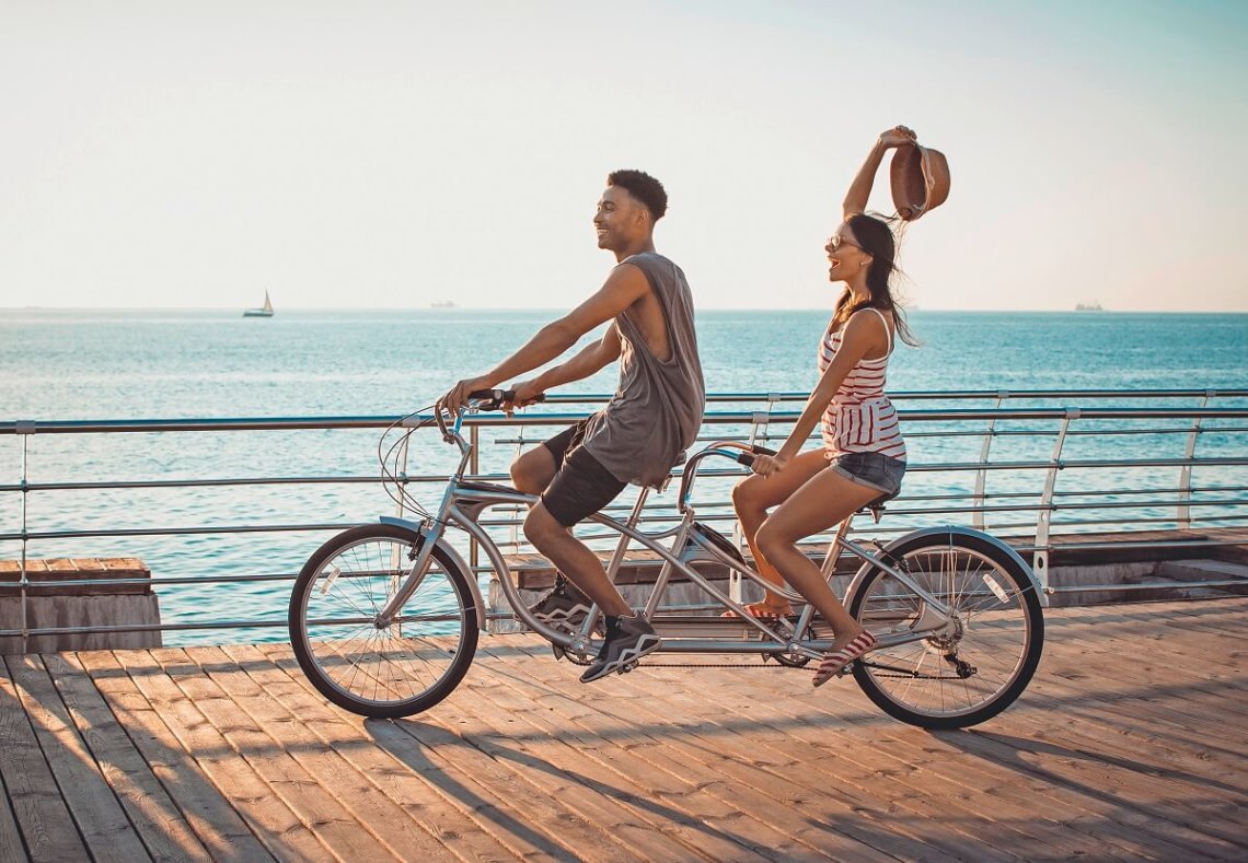 Symboldbild: Der Loop als Tandem-Partner im Diabetes-Alltag: Gemeinsam in die Pedale treten und lenken, aber auch mal loslassen dürfen. Ein Mann und eine Frau fahren Tandem in der Nähe des Meers.