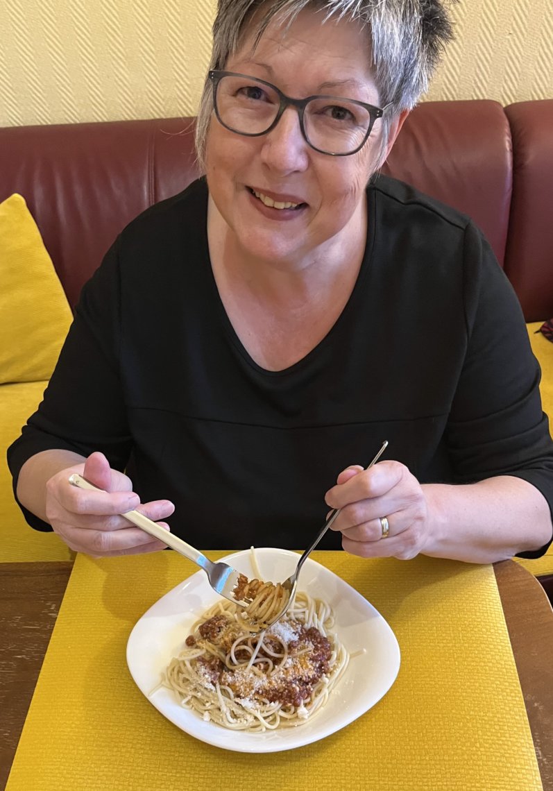 Susanne hat einen Teller mit Spaghetti Bolognese vor sich und hält das Besteck mit einigen Nudeln auf der Gabel