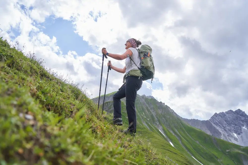 Conny wandert mit Stöcken und bepackt mit einem Rucksack eine Anhöhe hoch. Im Hintergrund ist ein Alpen-Panorama zu sehen.
