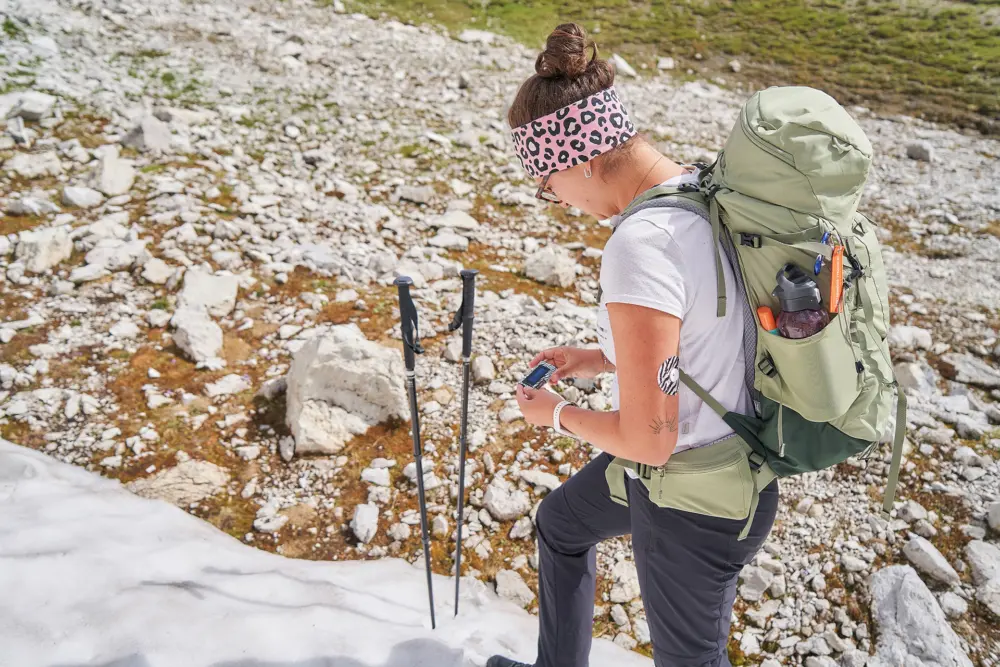 Conny mit großem Rucksack und einem CGM-Sensor am Arm prüft auf einem alpinen Geröllfeld die Einstellungen ihrer Insulinpumpe. Neben ihr stehen zwei Trekkingstöcke; im Vordergrund liegt ein Schneefeld.