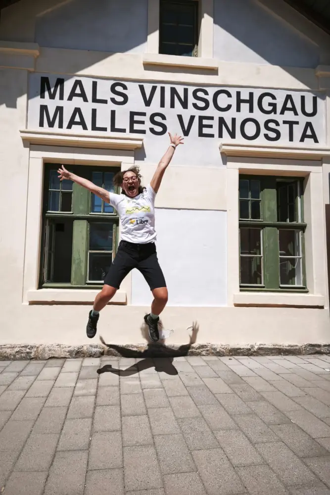 Conny springt mit ausgebreiteten Armen vor einem Gebäude mit der Aufschrift „MALS VINSCHGAU“ und „MALLES VENOSTA“ in die Luft. Sonnenschein wirft ihren Schatten auf den gepflasterten Boden.