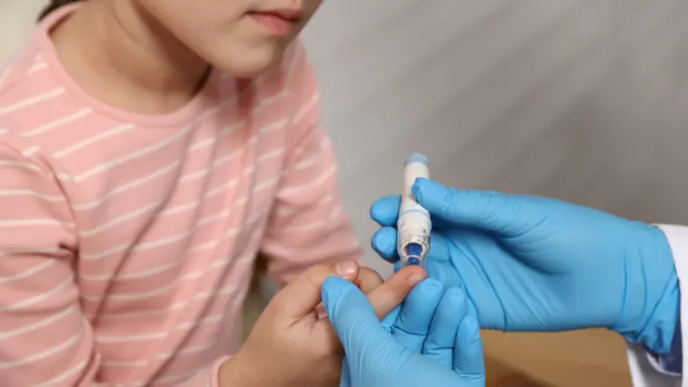 Eine medizinische Fachkraft mit blauen Einmalhandschuhen nutzt an einem Finger eines Mädchens in rosafarbenem Ringelshirt eine Stechhilfe, um Blut für die Glukosemessung zu gewinnen.