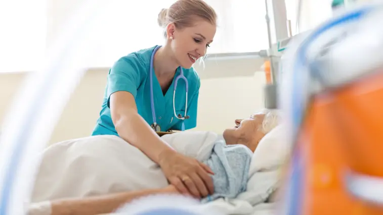 Eine junge Krankenschwester kümmert sich um eine ältere Frau in einem Krankenhausbett. Beide lächeln sich an.