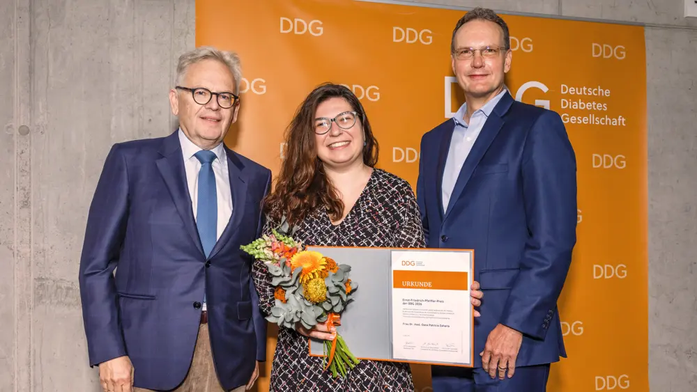 PD Dr. Oana-Patricia Zaharia hält gemeinsam mit Prof. Dr. Michael Roden (links) und Dr. Alexander Horn eine Urkunde zum Ernst-Friedrich-Pfeiffer-Preis der Deutschen Diabetes Gesellschaft. Sie trägt einen Blumenstrauß; im Hintergrund ist das orangefarbene DDG-Logo zu sehen.