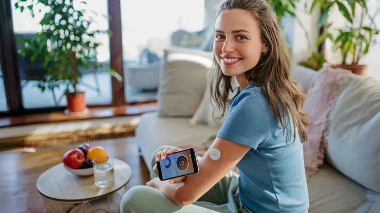 Junge Frau mit Glukosesensor am Oberarm hält ein Smartphone mit geöffneter Diabetes-App in den Händen und lächelt in die Kamera. Dargestellt wird die digitale Blutzuckerkontrolle im Alltag.