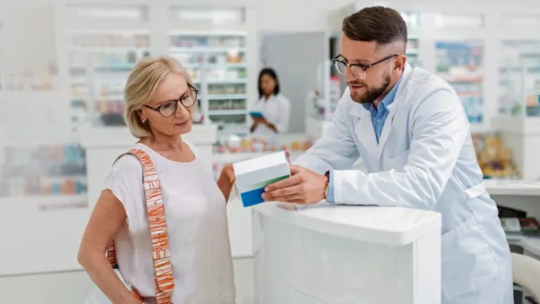 Apotheker erklärt einer älteren Kundin an der Ladentheke einer Apotheke eine Medikamentenpackung. Im Hintergrund sind Regale mit weiteren Arzneimitteln und eine Mitarbeiterin zu sehen.