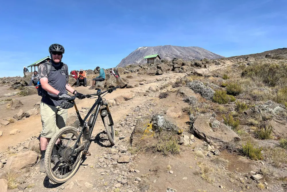 Markus Wielens mit Helm steht mit seinem Mountainbike auf einem steinigen Weg am Kilimandscharo. Im Hintergrund sind ein Bergmassiv, Schutzhütten und weitere Tourteilnehmende zu sehen.