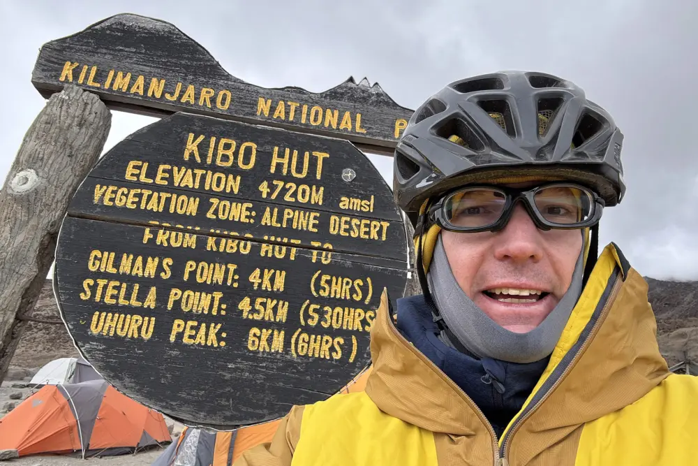 Markus Wielens in gelber Jacke und Helm posiert neben einem Wegweiser zur Kibo Hut auf 4.720 Metern Höhe am Kilimandscharo.
