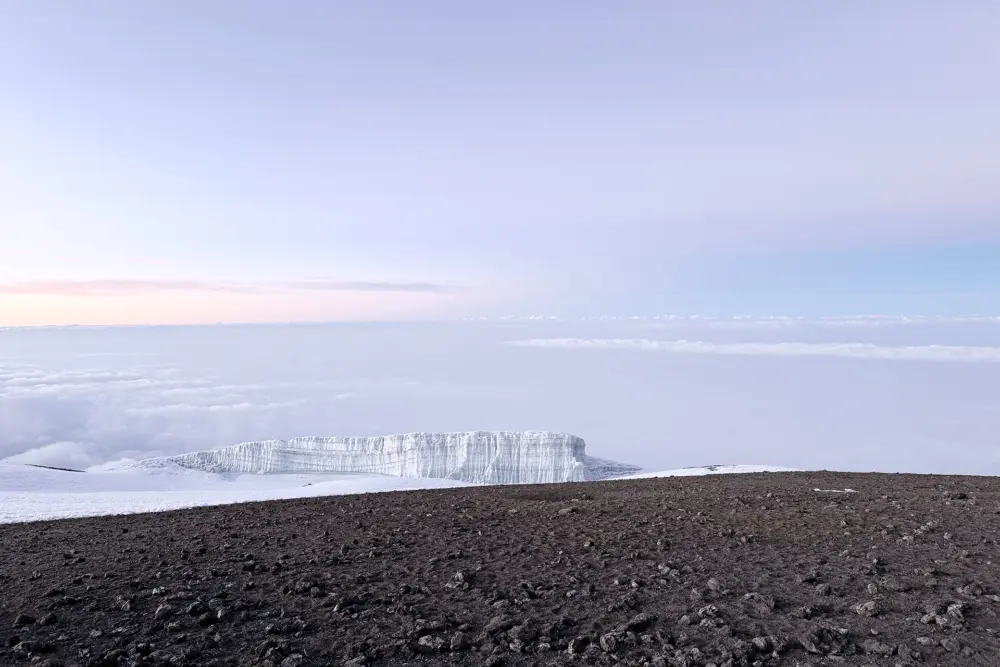 Verschneite Gletscherlandschaft am Kilimandscharo unter einem hellen, wolkenverhangenen Himmel und der aufgehenden Sonne im Hintergrund.