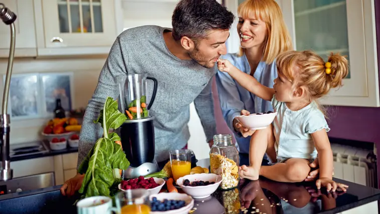 Eine Familie bereitet in der Küche gemeinsam ein ballaststoffreiches Frühstück zu. Ein Mann mixt grünes Blattgemüse und Karotten, während eine Frau und ein Mädchen mit Schalen voller Joghurt, Beeren und Haferflocken danebenstehen.