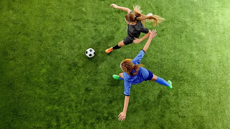 Zwei Fußballspielerinnen in schwarzen und blauen Trikots laufen auf einem grünen Rasen einem Fußball hinterher. Die Aufnahme aus der Vogelperspektive symbolisiert Bewegung und Prävention.