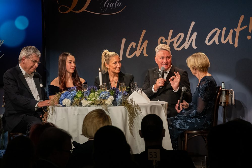 Anna Fernstädt, Janine Kunze und Hubert Fella sitzen mit Dr. Jens Kröger und Nicole Mattig-Fabian an einem blumengeschmückten Tisch auf der Bühne unter dem Schriftzug „Ich steh auf!“, Fella spricht gerade ins Mikrofon.