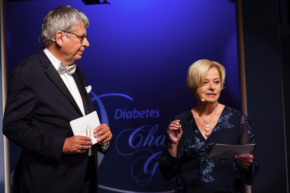 Dr. Jens Kröger und Nicole Mattig-Fabian moderieren auf der Bühne der Diabetes-Charity-Gala. Kröger hält Unterlagen und blickt zu Mattig-Fabian, die ins Mikrofon spricht und ihre Moderationskarten hält.