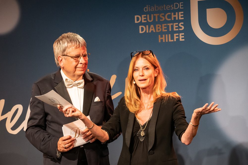 Esther Schweins spricht auf der Bühne und gestikuliert mit ausgebreiteten Armen. Neben ihr steht Moderator Dr. Jens Kröger mit Unterlagen in der Hand. Im Hintergrund ist das Logo von diabetesDE zu sehen.