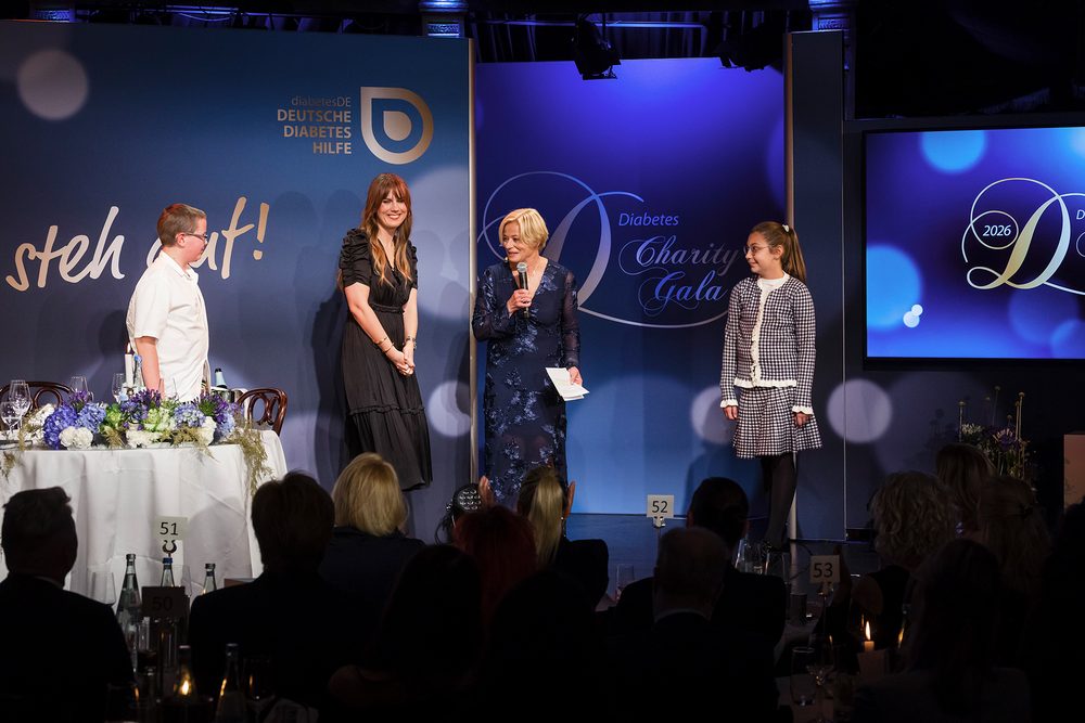 Isabelle Weberling steht mit zwei DiaConnect-Schützlingen und der Moderatorin auf der Bühne der Diabetes-Charity-Gala unter dem Schriftzug „Ich steh auf!“.