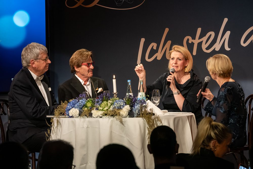 Jörg Draeger und Anke Harnack sitzen auf der Bühne an einem blumengeschmückten Tisch unter dem Schriftzug „Ich steh auf!“ und sprechen ins Mikrofon, neben ihnen Dr. Jens Kröger und Nicole Mattig-Fabian.