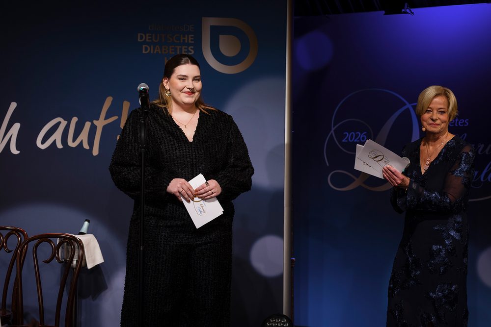 Lina Patti steht mit Notizen am Mikrofon auf der Bühne. Neben ihr hält eine Moderatorin einen Umschlag in den Händen.