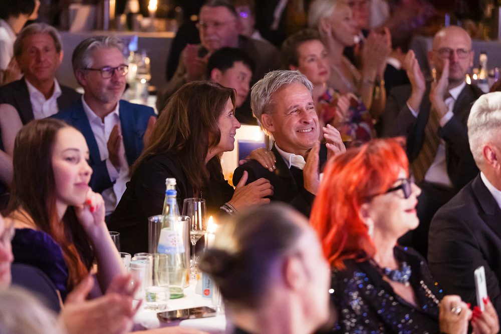 Pierre Littbarski applaudiert lächelnd inmitten des Publikums bei der Diabetes-Charity-Gala, neben ihm eine Begleiterin mit der Hand auf seiner Schulter.