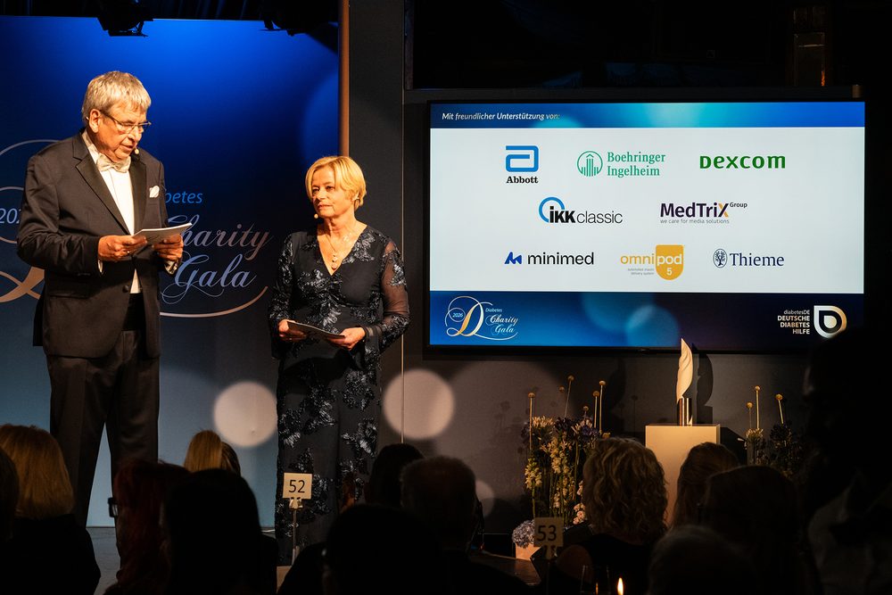 Dr. Jens Kröger und Nicole Mattig-Fabian stehen auf der Bühne neben einer Leinwand mit den Logos der unterstützenden Unternehmen Abbott, Boehringer Ingelheim, Dexcom, IKK classic, MedTriX Group, Minimed, Omnipod 5 und Thieme.
