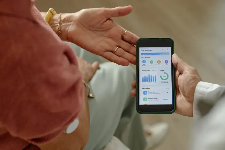 Eine Person mit Glukosesensor am Oberarm betrachtet gemeinsam mit einer zweiten Person die Diabetesdaten auf einem Smartphone. Die Anzeige zeigt Blutzuckerwerte, Tagesverlauf und dokumentierte Mahlzeiten.