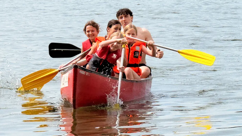 Fünf Kinder und Jugendliche mit orangen Schwimmwesten paddeln gemeinsam in einem roten Kanu auf dem Bodensee.