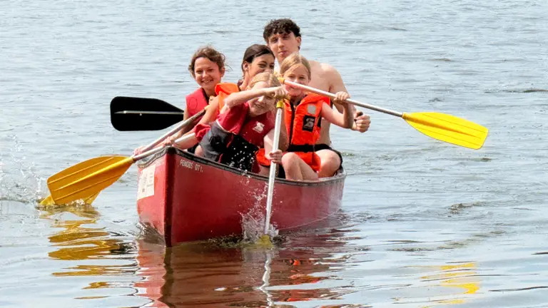 Fünf Kinder und Jugendliche mit orangen Schwimmwesten paddeln gemeinsam in einem roten Kanu auf dem Bodensee.