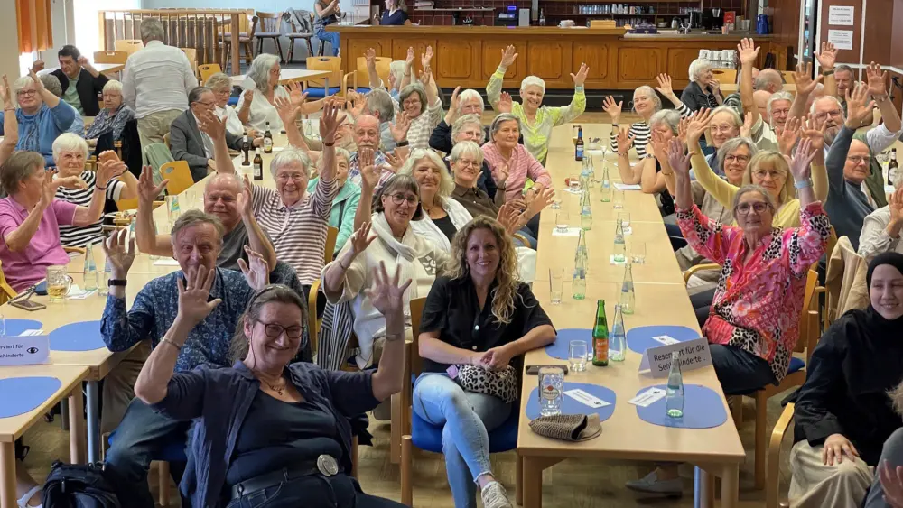 Teilnehmende des Hamburger Gehörlosen-Seniorenkreises sitzen an langen Tischen in einem Saal und winken für ein Gruppenfoto in die Kamera.