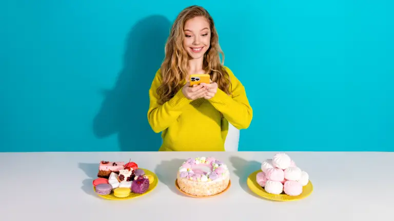 Junge Frau vor türkisem Hintergrund fotografiert mit ihrem Smartphone drei Teller mit Süßigkeiten, Kuchen und Gebäck, die vor ihr auf einem Tisch stehen.