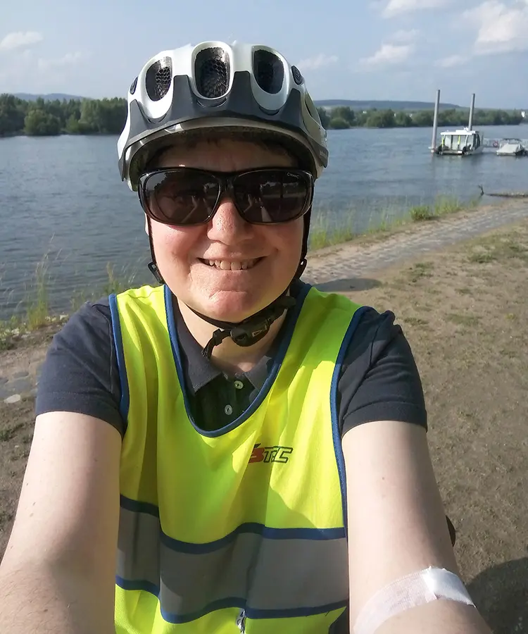 Selfie von Dr. med. Katrin Kraatz mit Fahrradhelm, Sonnenbrille und gelber Warnweste, im Hintergrund ein Fluss mit Booten und bewaldetem Ufer.