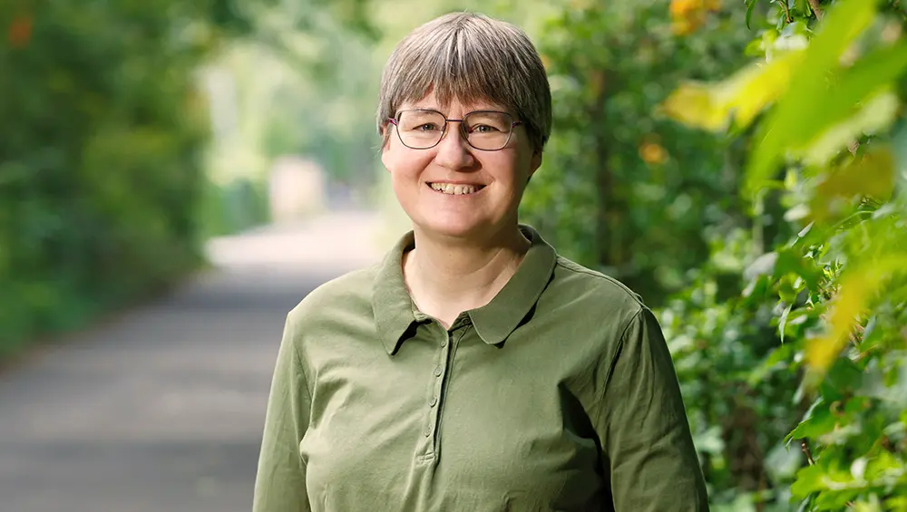 Porträt von Dr. med. Katrin Kraatz, sie trägt ein grünes Poloshirt und eine Brille und steht lächelnd auf einem Weg zwischen grünen Hecken und Bäumen.