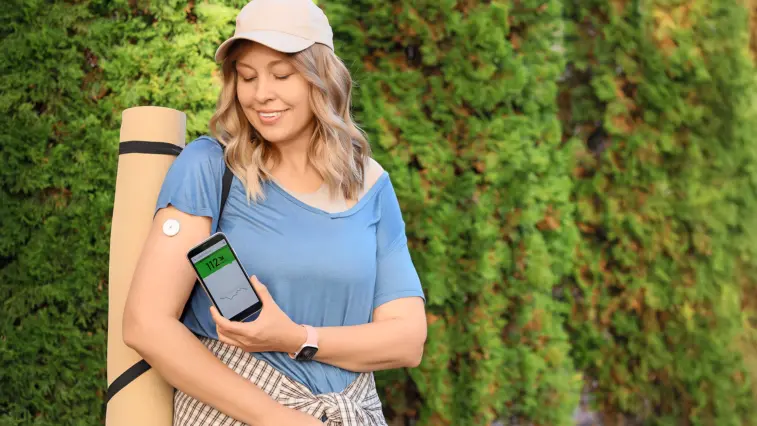 Eine Frau mit Rucksack und Yogamatte steht vor einer grünen Hecke. Sie trägt einen Glukosesensor am Oberarm und betrachtet auf ihrem Smartphone einen Glukosewert mit Verlaufskurve.