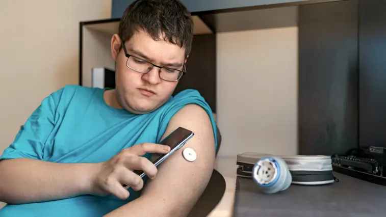 Ein Jugendlicher mit Brille betrachtet auf einem Smartphone seinen Glukosesensor am Oberarm. Auf dem Tisch liegen weitere Diabetes-Utensilien.