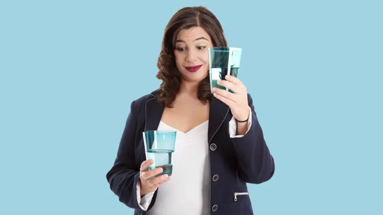 Eine Frau vergleicht zwei Gläser mit unterschiedlich hohem Wasserstand und blickt dabei nachdenklich auf das fast volle Glas.