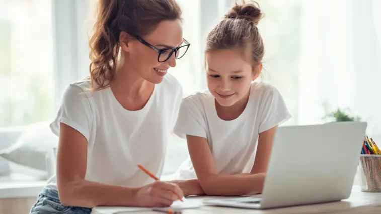 Eine Frau mit Brille und ein Mädchen sitzen gemeinsam an einem Tisch vor einem Laptop. Die Frau macht sich Notizen, während beide auf den Bildschirm schauen.