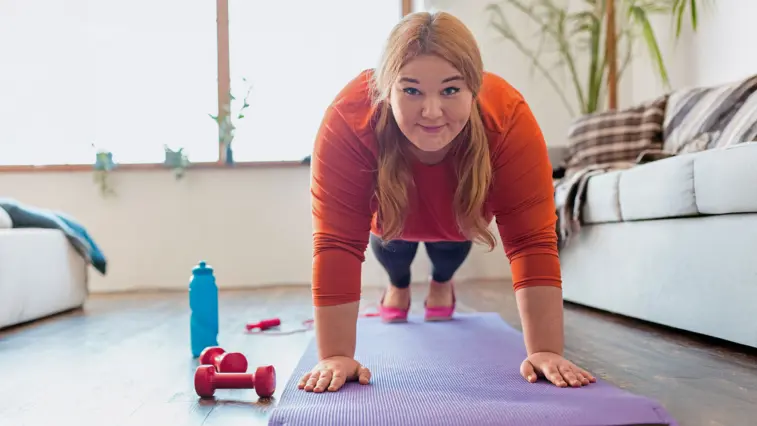 Eine Frau macht auf einer Trainingsmatte eine Plank-Übung in einem Wohnzimmer. Neben ihr liegen rote Hanteln und eine blaue Trinkflasche.