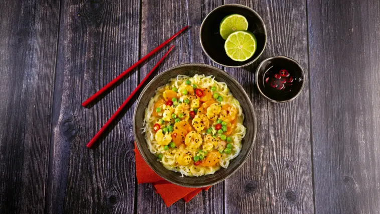Curry-Suppe mit Garnelen, Glasnudeln, Möhren, Erbsen und Chili in einer Schale auf einem dunklen Holztisch; daneben liegen rote Essstäbchen sowie Schälchen mit Limetten und Chili.