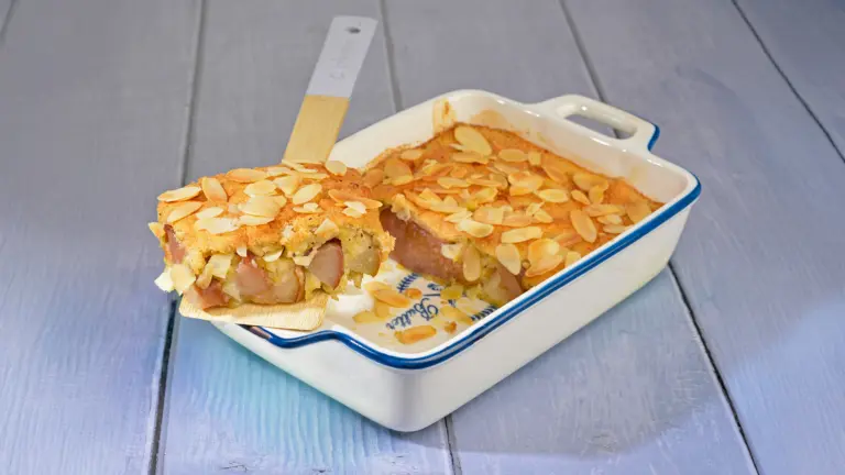 Mandel-Birnen-Auflauf in einer weiß-blauen Auflaufform, goldbraun gebacken und mit Mandelblättchen bestreut, ein angeschnittenes Stück mit sichtbaren Birnenstücken liegt auf einem Holzspatel.