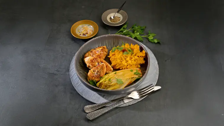 Gebratene Putenfilets mit Kürbispüree und gebratenem Chicorée, mit Petersilie bestreut, in einer Schale auf einem dunklen Tisch angerichtet. Daneben liegen Besteck und kleine Schälchen mit Gewürzen.