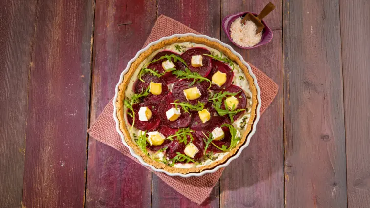 Rote-Bete-Tarte in einer weißen Tarte-Form, belegt mit dünnen Rote-Bete-Scheiben, Camembert-Stücken und Rucola, auf einem rosa Tuch und einem Holztisch, daneben eine kleine lila Schale mit grobem Salz.