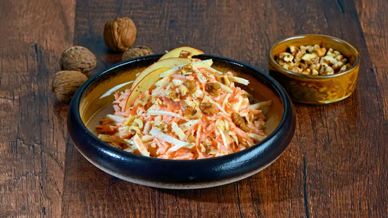 Sellerie-Möhren-Salat mit Apfel und gehackten Walnüssen in einer Schale auf einem Holztisch; daneben steht ein Schälchen mit weiteren Walnüssen.
