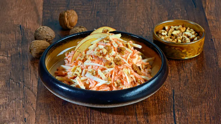 Sellerie-Möhren-Salat mit Apfel und gehackten Walnüssen in einer Schale auf einem Holztisch; daneben steht ein Schälchen mit weiteren Walnüssen.