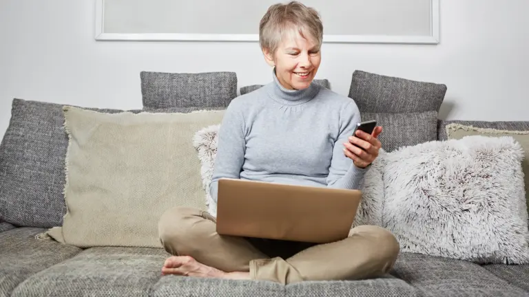 Eine lächelnde Frau sitzt mit einem Laptop auf dem Schoß auf einem Sofa und schaut auf ihr Smartphone.