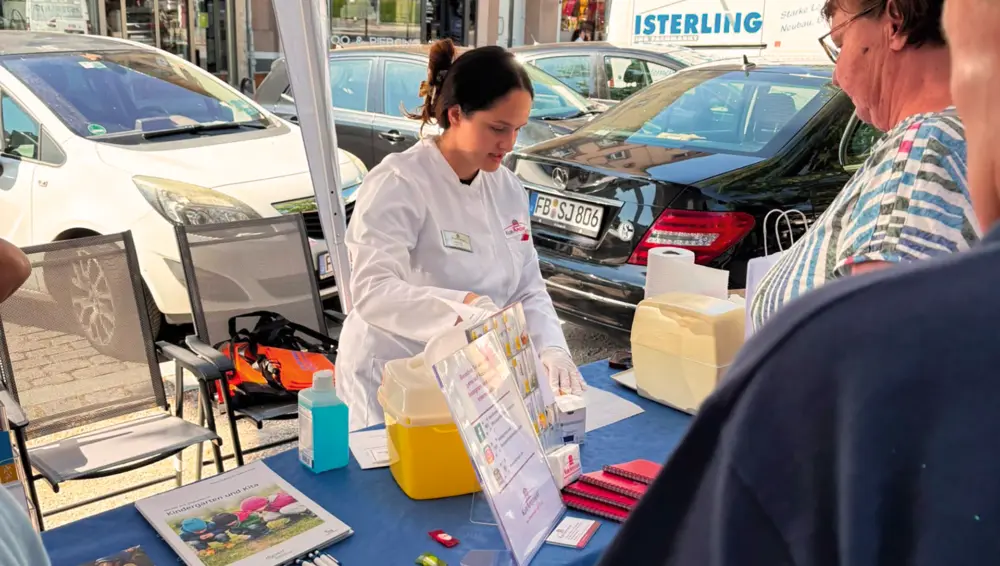 An einem Informationsstand unter einem Pavillon berät eine Frau im weißen Kittel eine Besucherin. Auf dem Tisch liegen Broschüren und Materialien zur Blutzuckermessung.