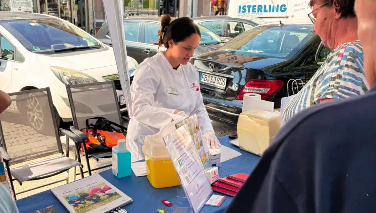 An einem Informationsstand unter einem Pavillon berät eine Frau im weißen Kittel eine Besucherin. Auf dem Tisch liegen Broschüren und Materialien zur Blutzuckermessung.