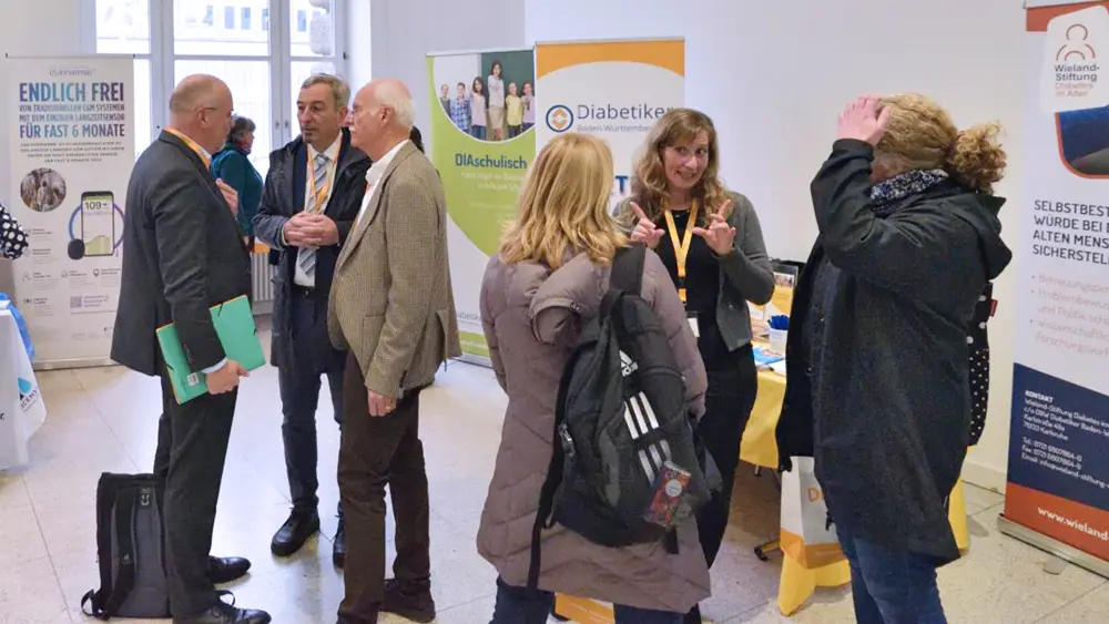 Mehrere Personen im Gespräch an Informationsständen bei einer Diabetes-Veranstaltung, im Hintergrund Roll-ups unter anderem der Diabetiker Baden-Württemberg und der Wieland-Stiftung.