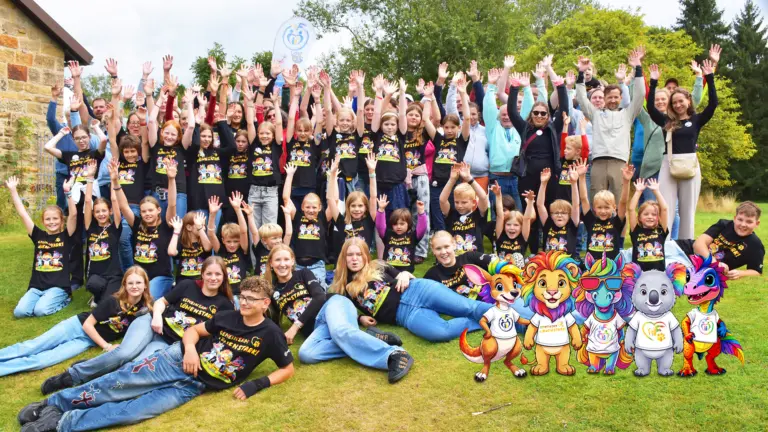Großes Gruppenfoto auf einer Wiese: Zahlreiche Kinder, Jugendliche und Eltern in schwarzen T-Shirts mit der Aufschrift „Gemeinsam löwenstark!“ strecken jubelnd die Arme in die Höhe, rechts unten sind bunte Figuren von Fantasietieren eingeblendet.
