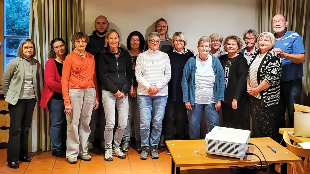 Gruppenfoto von 15 Personen in einem Vereinsraum nach einem Vortrag, im Vordergrund ein Beamer auf einem Tisch. Referentin Cornelia Sichermann steht als vierte Person von rechts in der Gruppe.