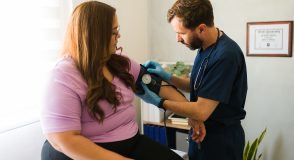 Arzt in dunkelblauem Kittel misst bei einer Patientin mit korpulenter Statur mit einem manuellen Blutdruckmessgerät den Blutdruck am Oberarm.